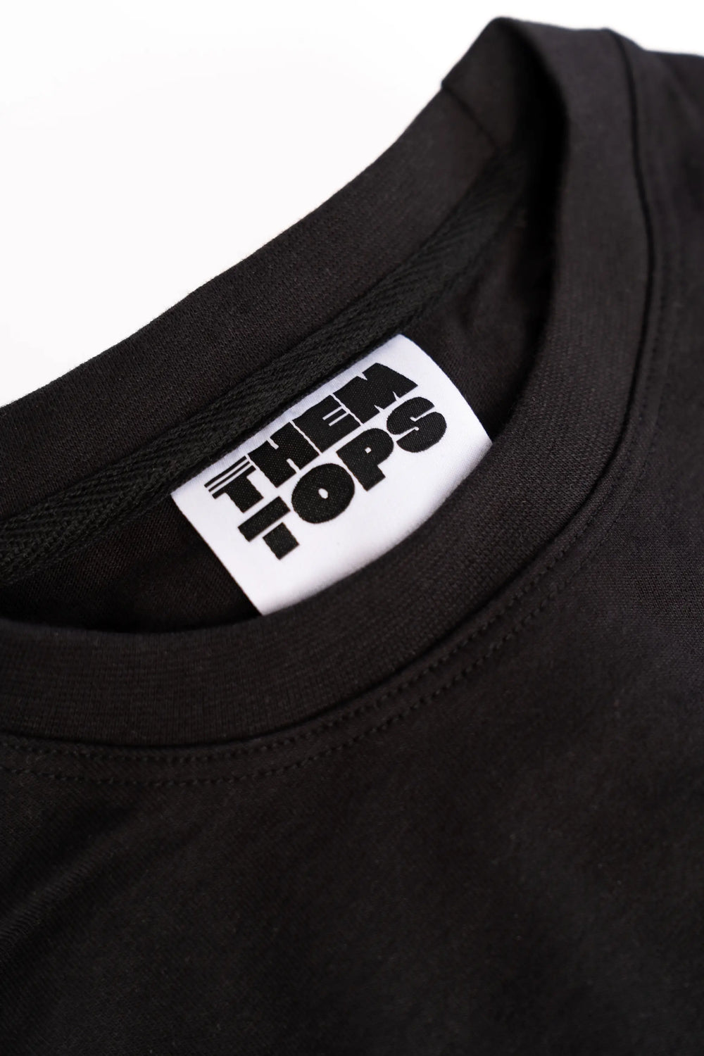 Close-up of a black crewneck t-shirt showing a white neck label with the brand name 'THEM TOPS' printed in black.