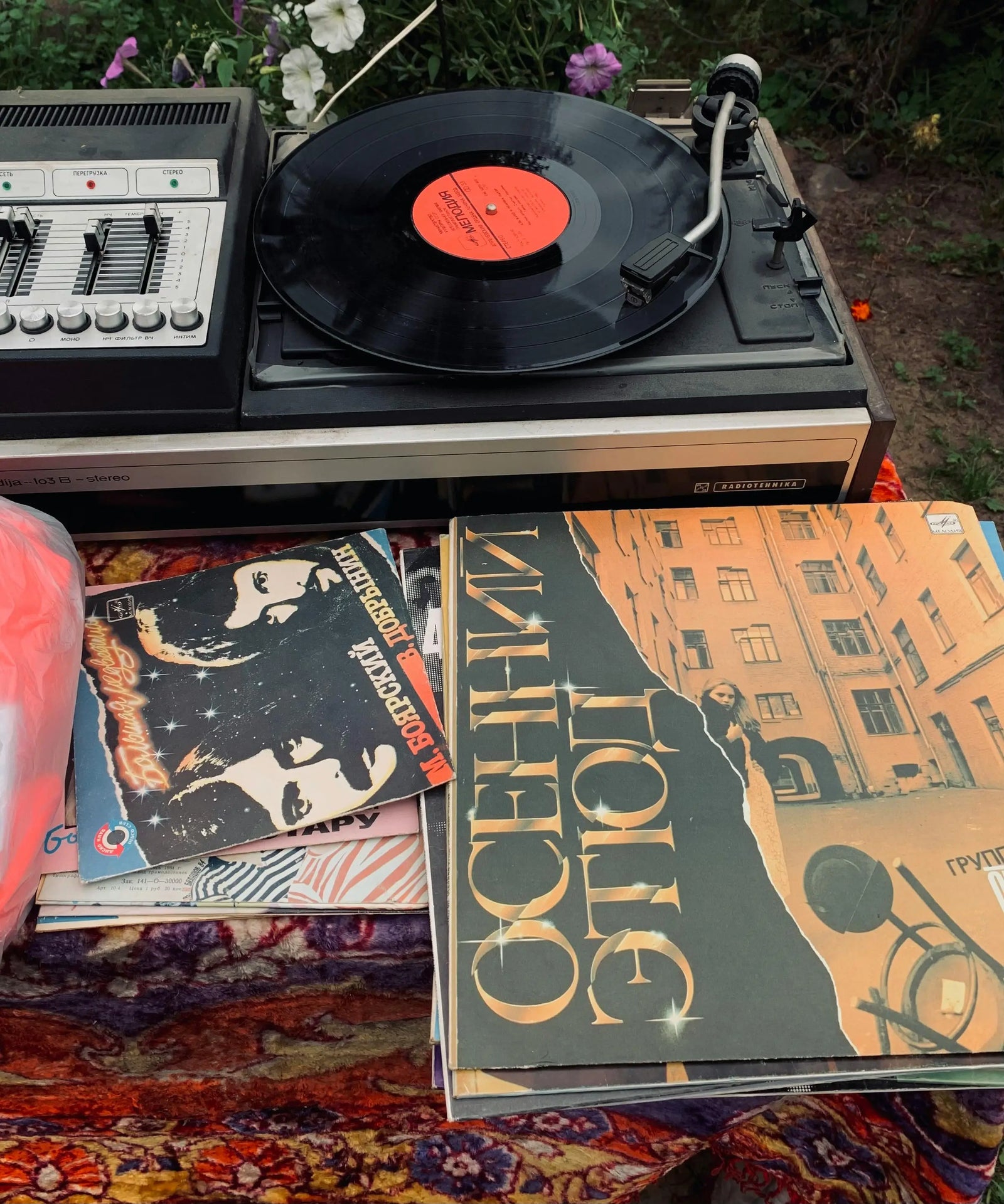 A vintage black turntable with a vinyl record spinning, set outdoors on top of stacked vinyl records. 