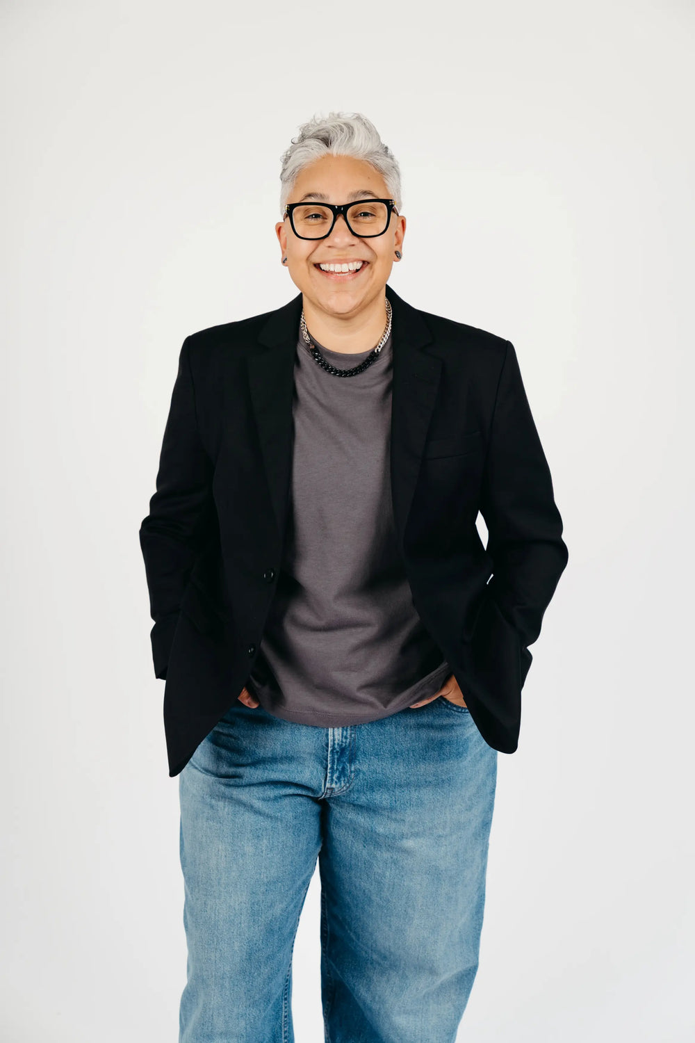 A smiling person with short gray hair and black glasses, wearing a black blazer over a dark gray t-shirt and blue jeans, standing against a plain white studio background.