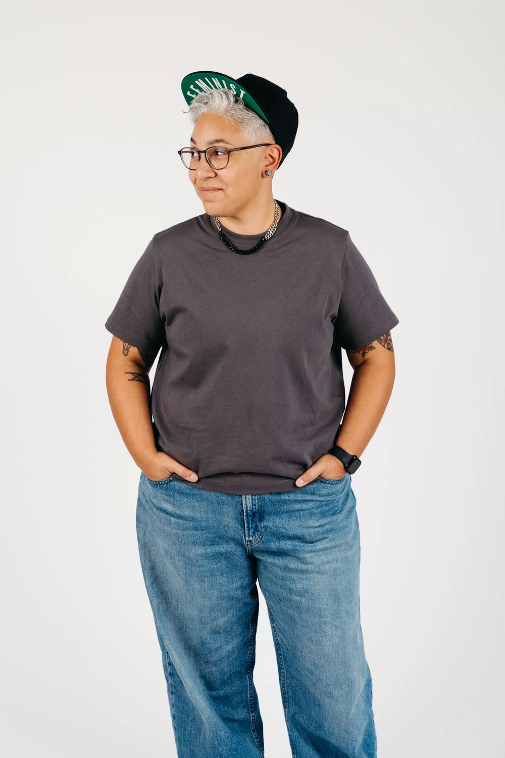 A person wearing a dark charcoal gray crewneck t-shirt, blue jeans, a black hat with a green and white pattern, and glasses, standing against a plain white studio background.
