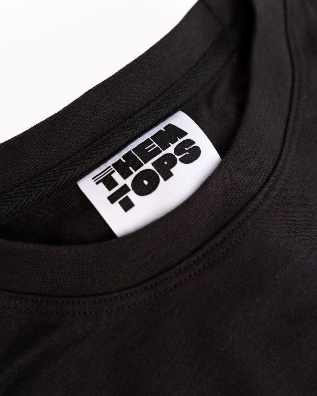 Close-up of a black crewneck t-shirt showing a white neck label with the brand name 'THEM TOPS' printed in bold black letters.