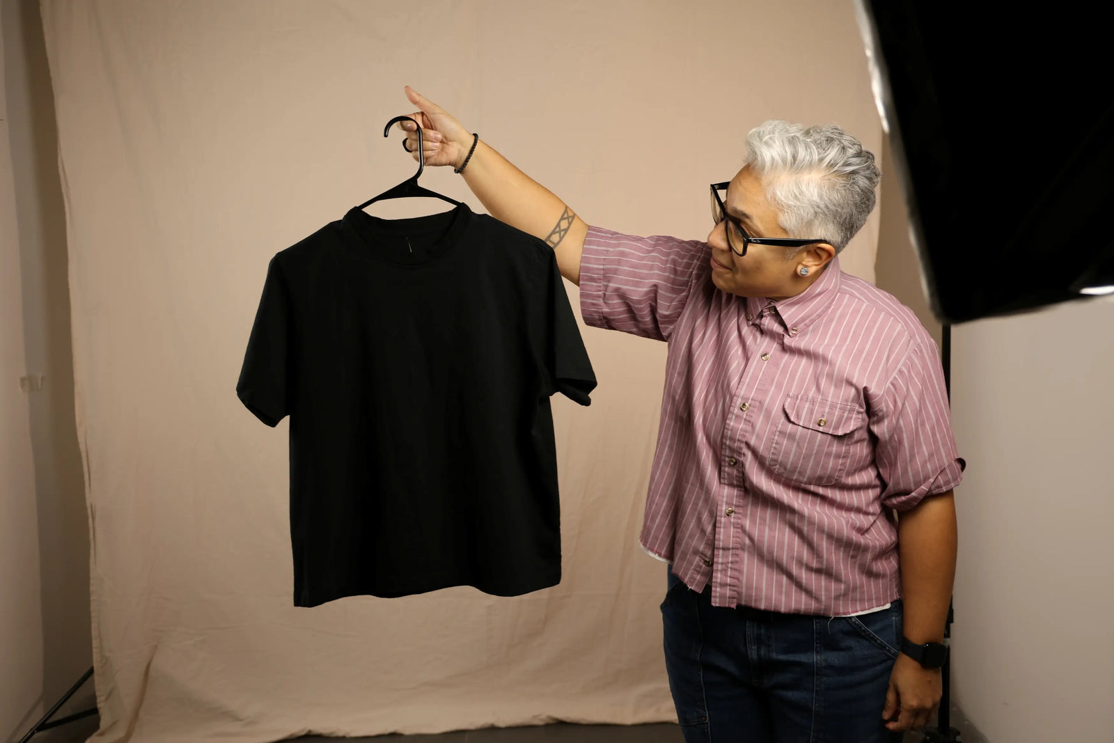 A person with short gray hair and glasses holds up a plain black short-sleeve t-shirt on a hanger against a beige fabric backdrop. The person is wearing a pink and white striped button-down shirt and dark jeans.