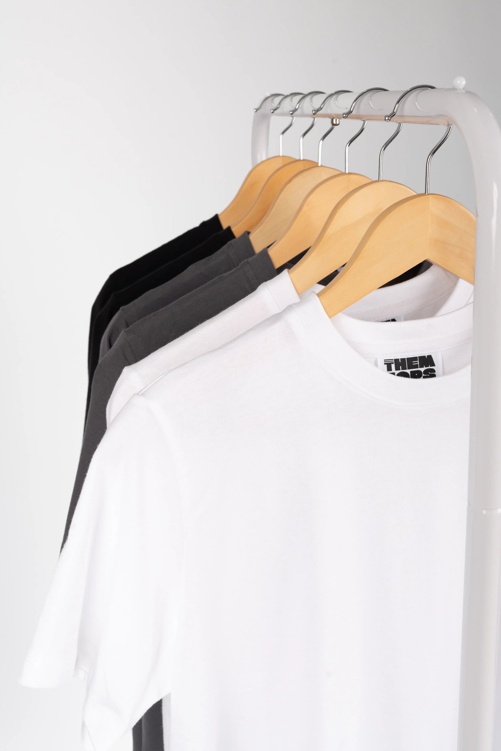 A white short-sleeve crewneck t-shirt hanging on a wooden hanger from a clothing rack, with black and gray t-shirts visible behind it.