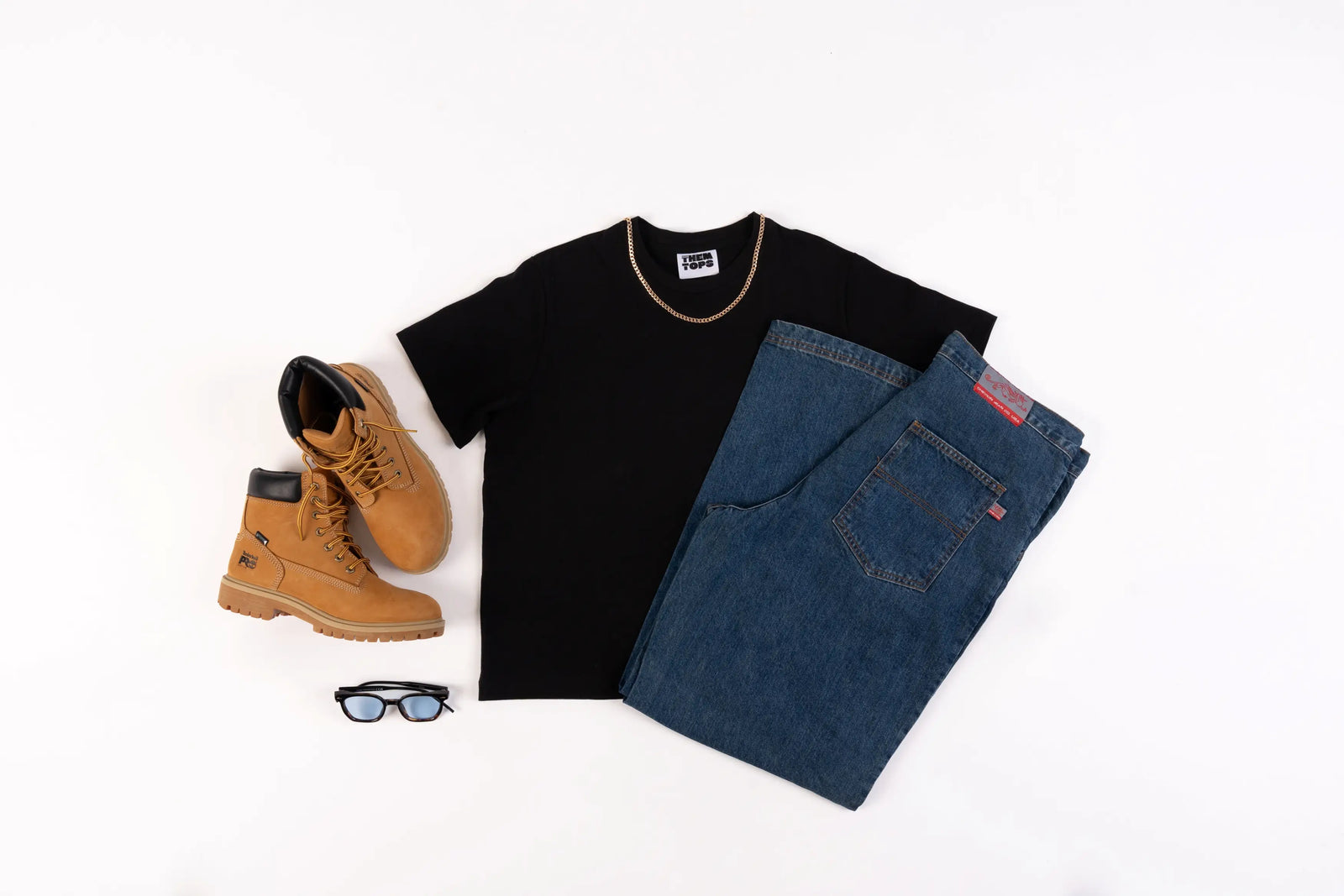 A styled flat-lay outfit on a white background featuring a black short-sleeve t-shirt, blue jeans, tan lace-up boots, and a pair of black sunglasses.