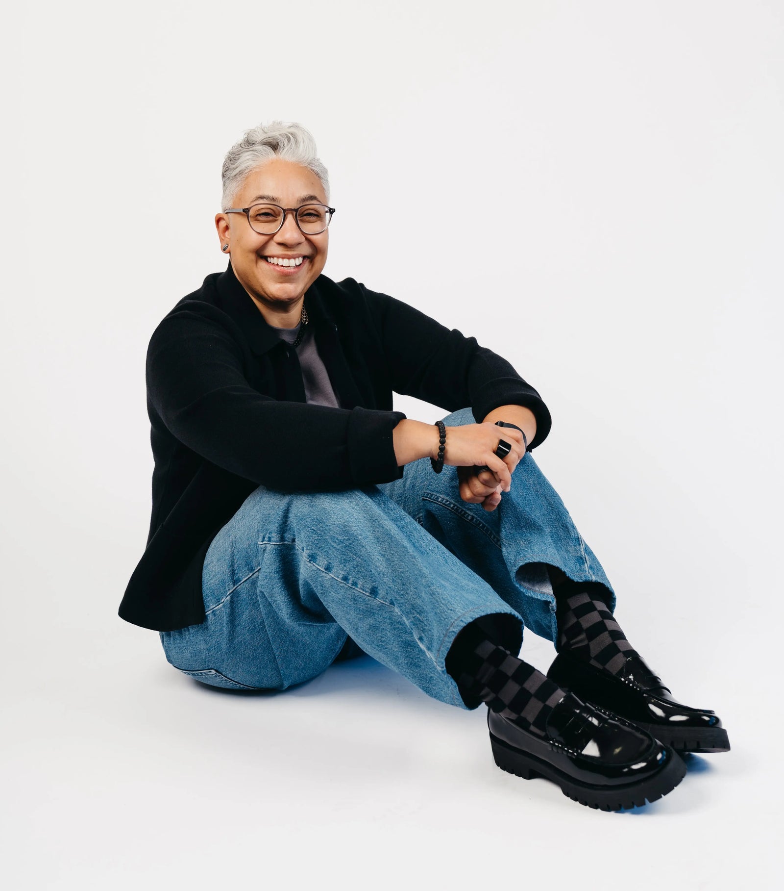 A smiling person with short gray hair and glasses sits on a white studio background, wearing a black cardigan, blue jeans, and black patent loafers.
