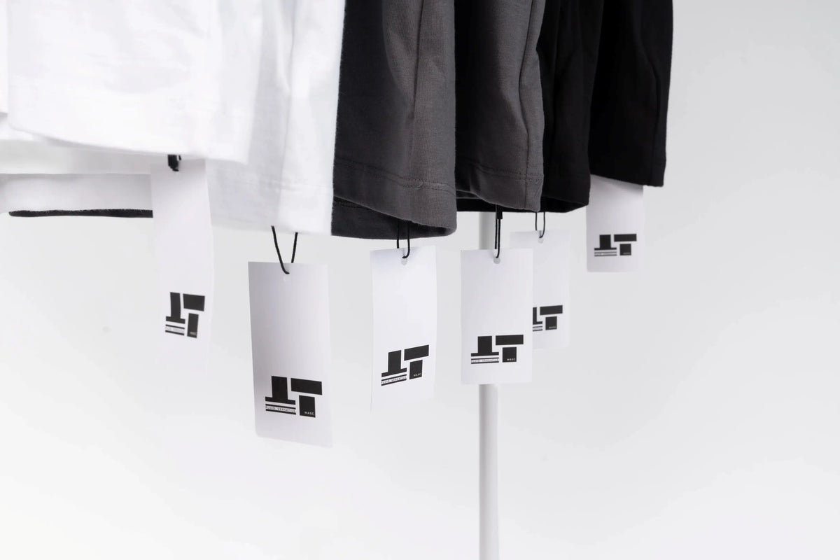 A close-up of hanging garments in white, dark gray, and black, each with a white tag featuring a black geometric logo, set against a clean white background.